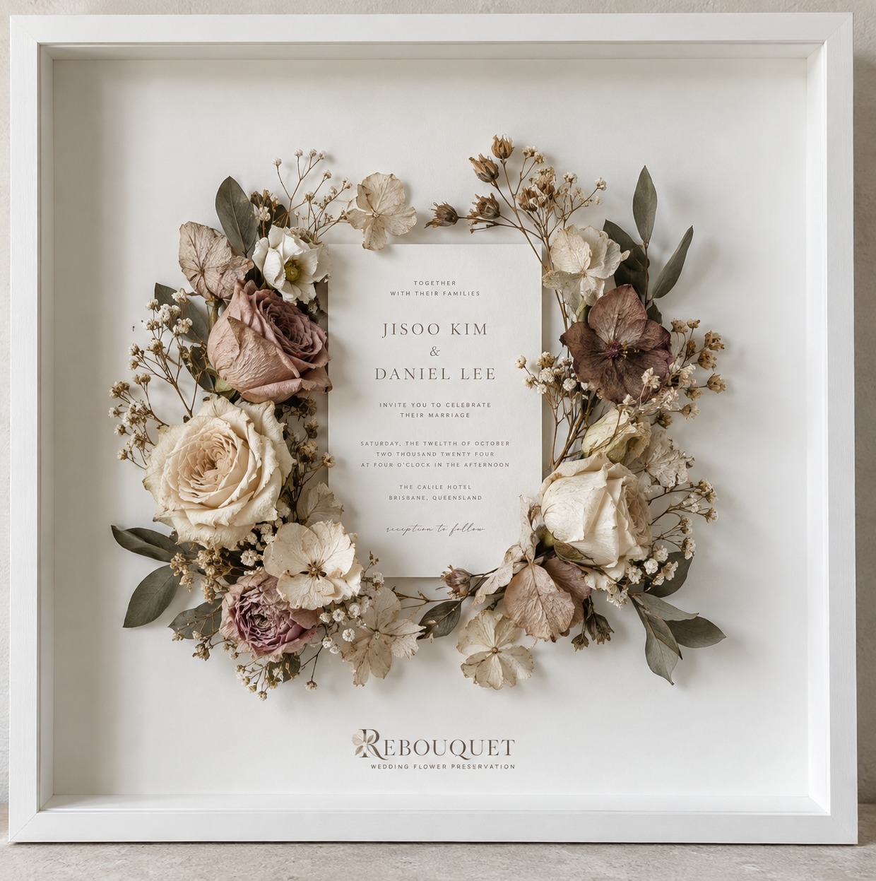 Preserved wedding invitation surrounded by dried flower wreath in a white shadow box frame